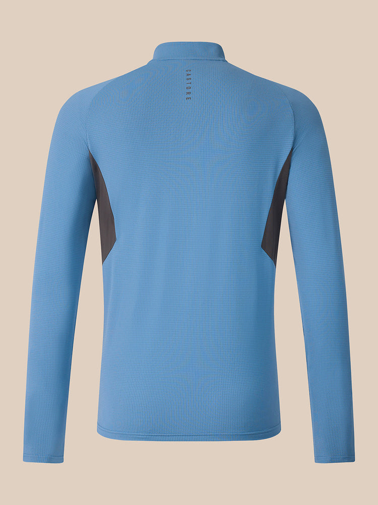 A light blue long-sleeve athletic quarter-zip pullover shown from the back, featuring dark gray side panel accents and a small vertical brand logo near the neckline, set against a solid beige background.