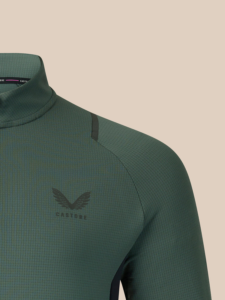Close-up of a dark green athletic quarter-zip top with a winged logo and the word 'CASTORE' on the left chest, set against a solid beige background.