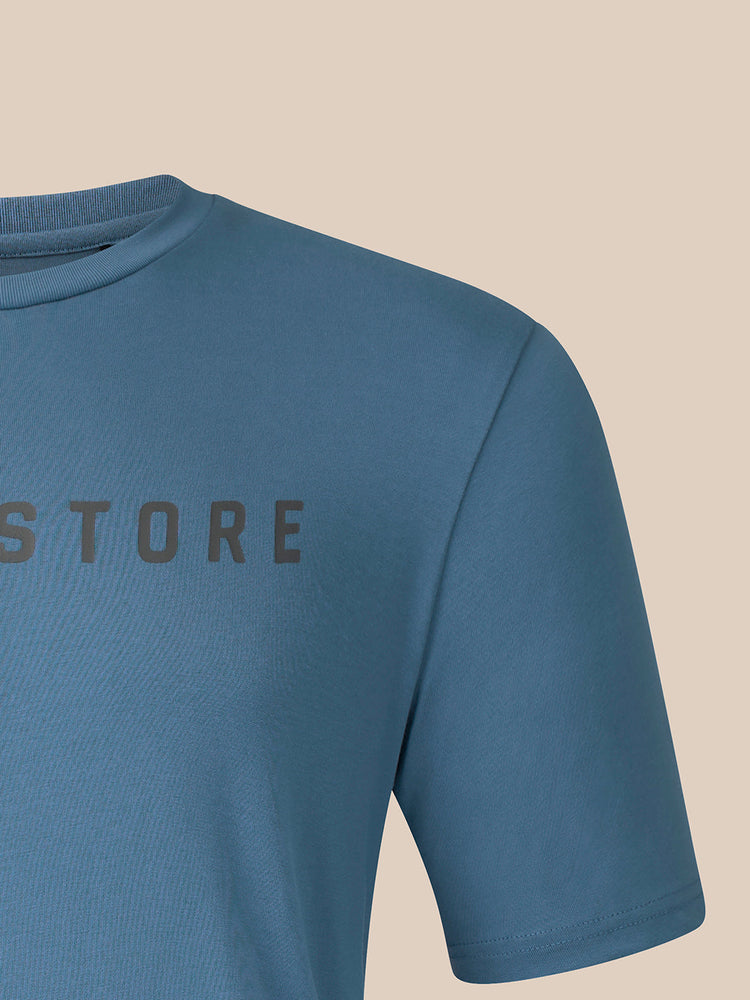 Close-up of a blue short-sleeve crewneck t-shirt with a dark gray 'CASTORE' text graphic on the chest, shown on a solid beige background.